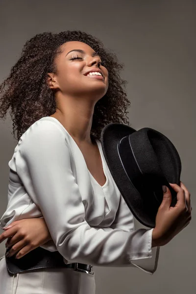 Stylish african american woman in white clothes smiling and waving black hat isolated on grey background — Stock Photo