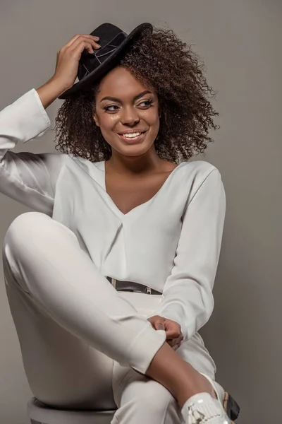Beautiful african american woman in white clothes smiling and holding black hat isolated on grey background — Stock Photo
