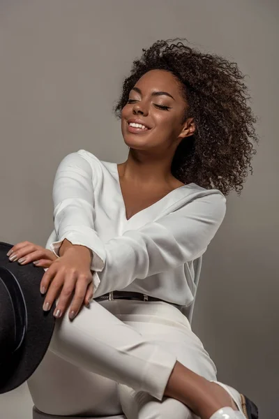 Young sensual african american woman in white clothes smiling and holding black hat isolated on grey background — Stock Photo