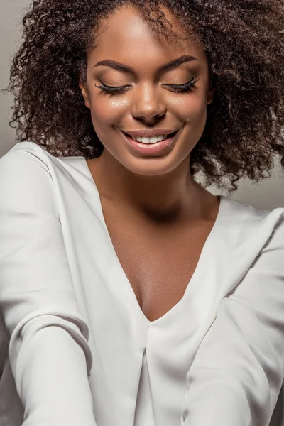 Elegante mujer afroamericana en camisa blanca sonriendo y mirando hacia abajo aislada sobre fondo gris - foto de stock