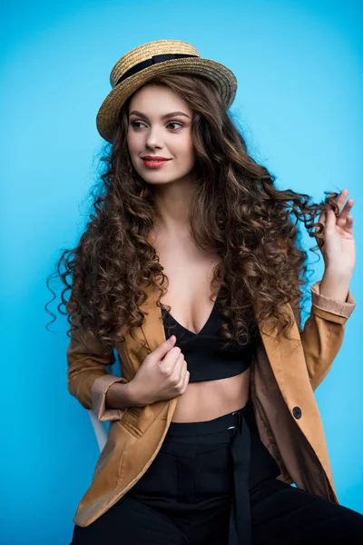 Attractive young woman with long curly hair in fashionable canotier hat and jacket isolated on blue — Stock Photo