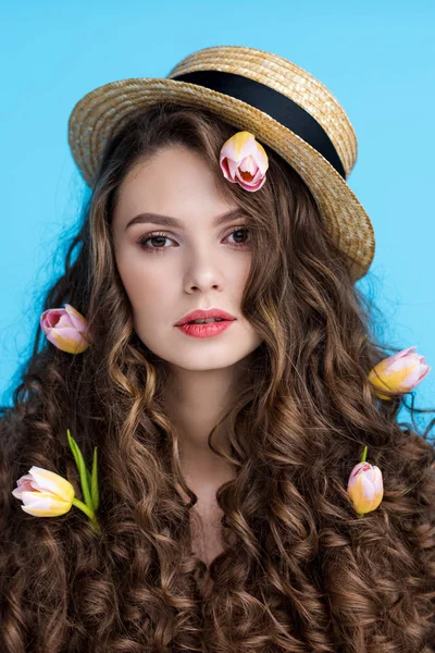 Sensual mujer joven en sombrero canotier con flores en su largo pelo rizado aislado en azul - foto de stock