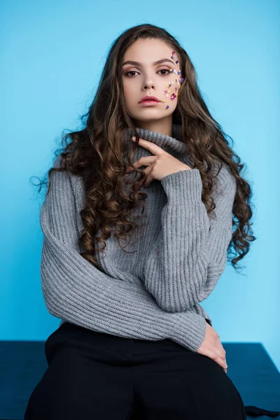 Élégant jeune femme aux cheveux longs avec des fleurs sur le visage en pull assis sur la table isolé sur le bleu — Photo de stock