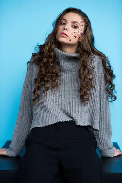Молода жінка з квітами на обличчі в стильному светрі, сидить на столі — стокове фото