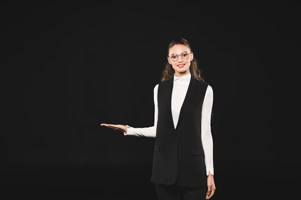 Beautiful brunette woman in eyeglasses showing copy space and smiling at camera isolated on black — Stock Photo