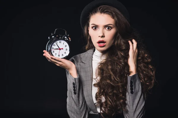 Shocked young woman holding clock and looking at camera isolated on black — Stock Photo