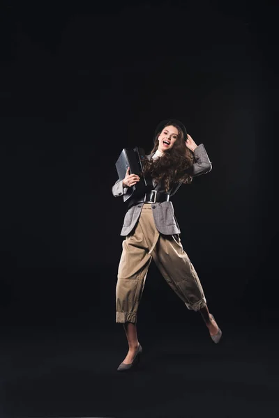 Alegre elegante mujer joven sosteniendo carpetas y mirando a la cámara aislada en negro - foto de stock