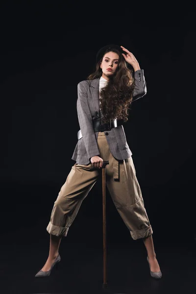 Stylish young woman posing with walking stick and looking at camera isolated on black — Stock Photo