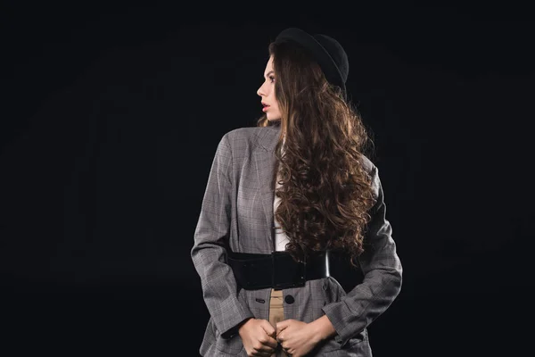 Beautiful brunette woman in jacket with black belt and hat looking away isolated on black — Stock Photo