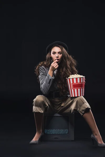 Шокированная молодая женщина ест попкорн, сидя на телевидении и глядя на камеру на черном — стоковое фото