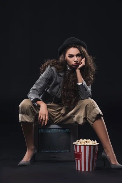 Серьезная стильная молодая женщина сидит на телевизоре с кукурузой и смотрит на камеру в черном — стоковое фото