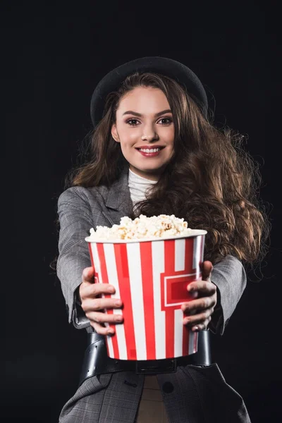 Hermosa mujer joven elegante sosteniendo palomitas de maíz y sonriendo a la cámara aislada en negro - foto de stock