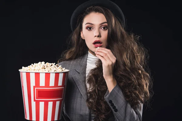 Schockierte modische junge Frau, die Popcorn isst und isoliert auf schwarz in die Kamera schaut — Stockfoto