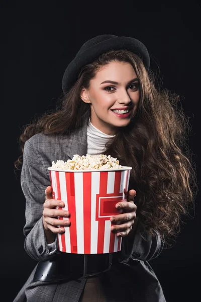 Hermosa mujer joven con estilo sosteniendo palomitas de maíz y sonriendo a la cámara aislada en negro - foto de stock