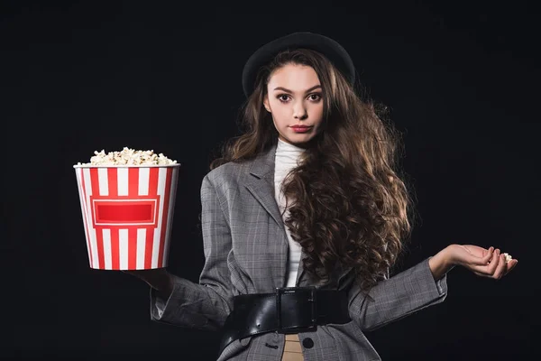 Beautiful stylish young woman holding popcorn and looking at camera isolated on black — Stock Photo