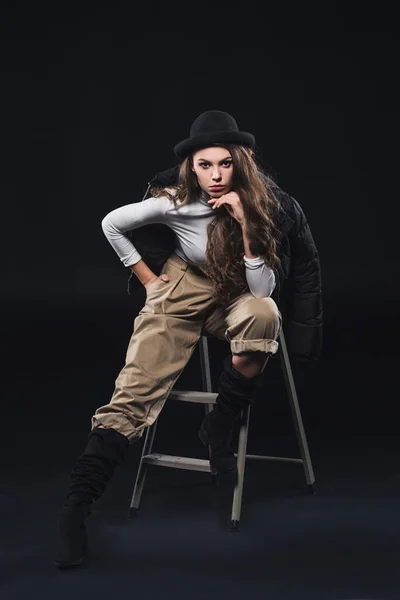 Beautiful young woman in fashionable hat and winter jacket sitting on ladder and looking at camera on black — Stock Photo