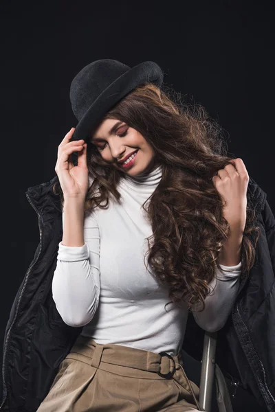 Улыбающаяся молодая женщина в стильной шляпе и зимней куртке сидит на лестнице изолированы на черном — стоковое фото