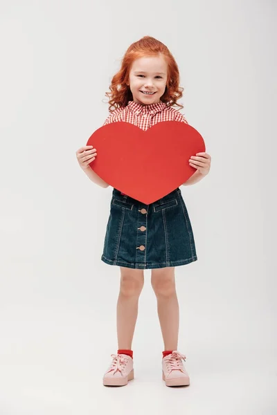 Bellissimo bambino che tiene il simbolo del cuore rosso e sorride alla fotocamera isolata sul grigio — Foto stock