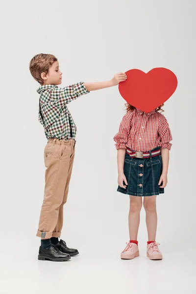 Niño pequeño sosteniendo el símbolo rojo del corazón cerca de la cara de niña aislada en gris - foto de stock