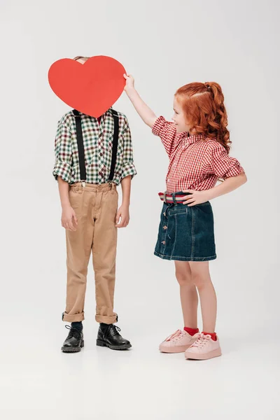 Niña sosteniendo el símbolo rojo del corazón cerca de la cara del niño aislado en gris - foto de stock