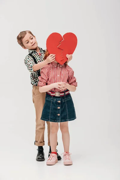 Niño pequeño sosteniendo el símbolo del corazón roto cerca de la cara de niña aislada en gris - foto de stock