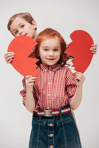 Little boy holding parts of broken heart and adorable little girl smiling at camera isolated on grey — Stock Photo