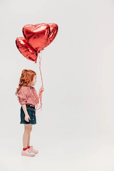 Очаровательный рыжий ребенок с красными шарами в форме сердца, изолированными на сером — стоковое фото