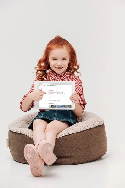 Adorável menina sorrindo para a câmera enquanto segurando tablet digital com site airbnb na tela isolada em cinza — Fotografia de Stock