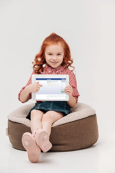 Adorable little girl smiling at camera while holding digital tablet with facebook website on screen isolated on grey — Stock Photo