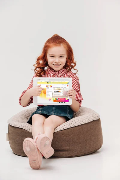 Entzückendes kleines Mädchen lächelt in die Kamera und hält ein digitales Tablet mit aliexpress-Website auf dem Bildschirm isoliert auf grau — Stockfoto