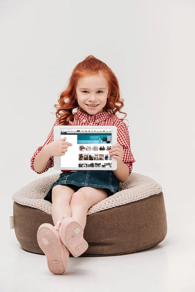Entzückendes kleines Mädchen lächelt in die Kamera, während es ein digitales Tablet mit Amazon-Website auf dem Bildschirm hält, isoliert auf grau — Stockfoto