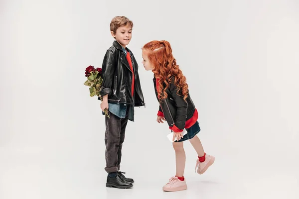 Niño ocultando rosas para su pequeña novia detrás de la espalda aislado en gris - foto de stock