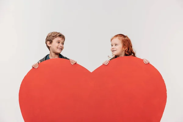 Smiling little kids couple behind large red heart isolated on grey — Stock Photo