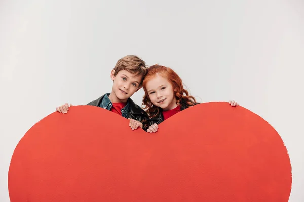 Happy bonding little kids behind large red heart isolated on grey — Stock Photo