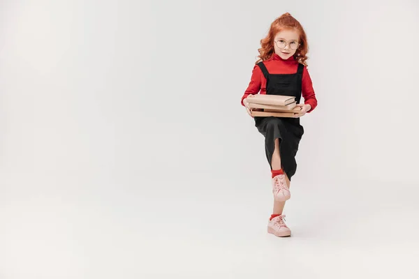 Entzückendes kleines Schulmädchen, das einen Stapel Bücher isoliert auf grau trägt — Stockfoto