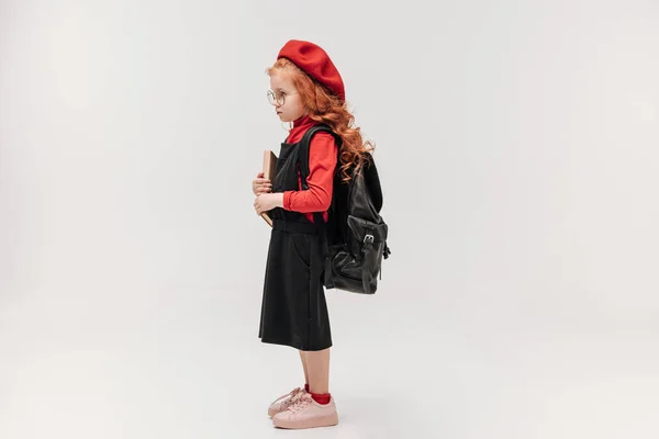 Vista laterale di adorabile scolaretta in berretto rosso con libro e zaino isolato su grigio — Foto stock