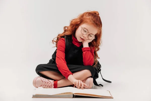 Adorable colegiala lectura libro mientras está sentado en el suelo aislado en gris - foto de stock