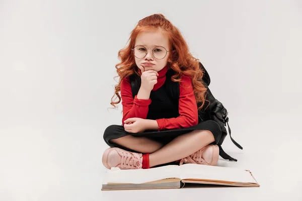 Скучающая маленькая школьница, сидящая на полу с открытой книгой, изолированной на сером — стоковое фото