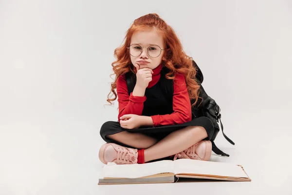 Pequeña colegiala aburrida sentada en el suelo con libro abierto aislado en gris - foto de stock