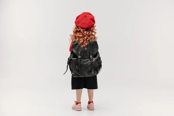 Посмотреть на маленькую школьницу с большим рюкзаком, изолированным на сером — стоковое фото