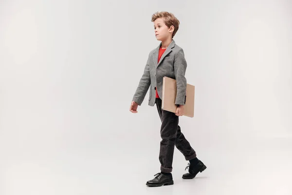 Elegante piccolo scolaro a piedi con grande libro isolato su grigio — Foto stock