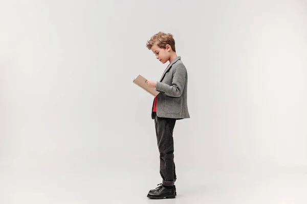 Side view of stylish little schoolboy reading book isolated on grey — Stock Photo