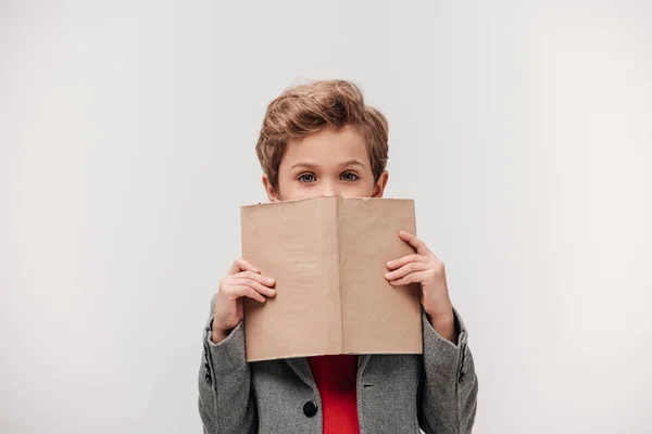 Стильний маленький школяр, що закриває обличчя з книгою ізольовано на сірому — стокове фото