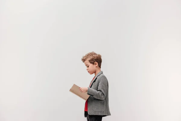 Vista laterale di elegante piccolo scolaro lettura libro isolato su grigio — Foto stock