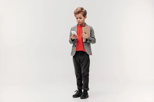 Élégant petit écolier avec smartphone et livre isolé sur gris — Photo de stock