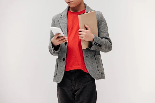 Schnappschuss eines stylischen kleinen Schülers mit Smartphone und Buch isoliert auf grau — Stockfoto