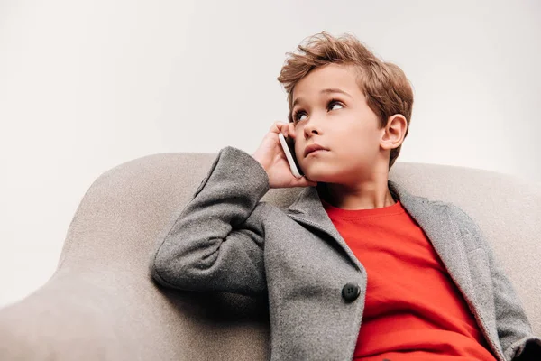 Niño pequeño y elegante hablando por teléfono mientras está sentado en sillón aislado en gris - foto de stock