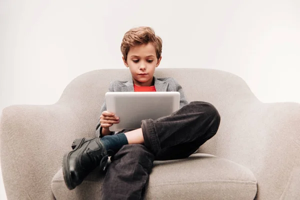 Serious little boy using tablet  while sitting in armchair isolated on grey — Stock Photo