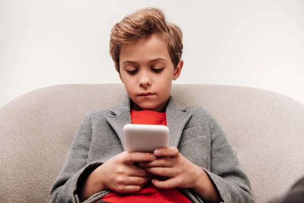 Niño serio usando el teléfono inteligente mientras está sentado en sillón aislado en gris - foto de stock