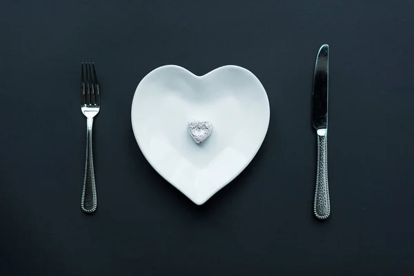 Vue de dessus de bonbons enveloppés sur plaque en forme de coeur blanc avec couverts isolés sur noir — Photo de stock